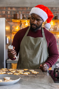 African American man icing cookies with piping bag on kitchen counter wearing Santa hat and apron