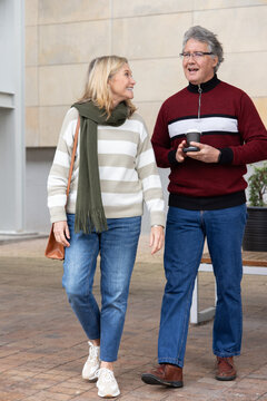 Senior couple walking and talking on paved plaza, holding takeaway coffee cup and shoulder bag