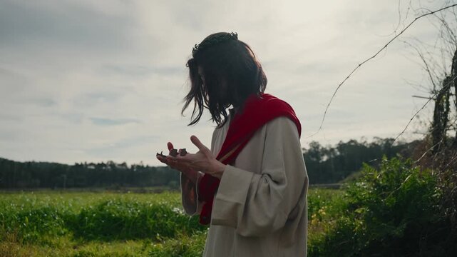 Actor portraying Jesus Christ in robe and red sash holding nails in wounded hands outdoors