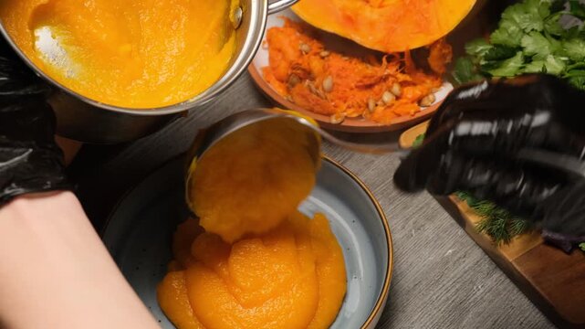 Chef hands in black gloves using ladle to serve hot vegetarian pumpkin porridge into bowl. Hot pumpkin puree as breakfast or dinner. Pumpkin soup puree. Vegan lifestyle