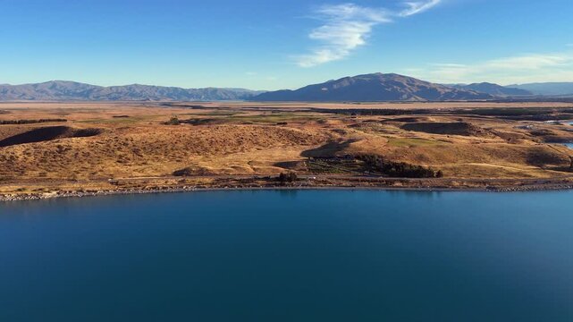 Flyover mountain shoreline to golden moraine meadow under blue sky