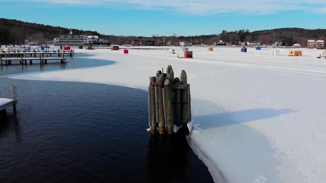 A partially frozen lake shows the edge of ice meeting open water, with colorful ice fishing shacks scattered across the snow-covered surface and wooden docks visible.