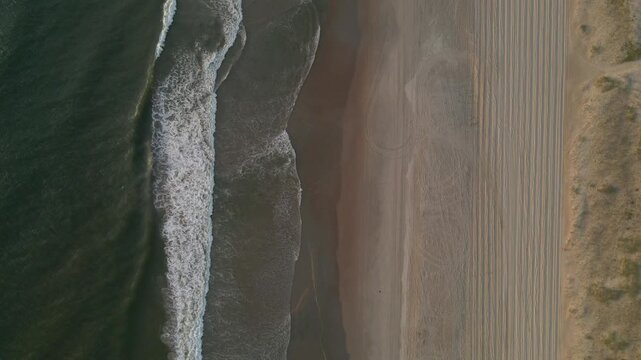 Drone footage captures the mesmerizing pattern of white ocean waves rolling onto a wide sandy beach with visible tire tracks and coastal vegetation in the Outer Banks North Carolina