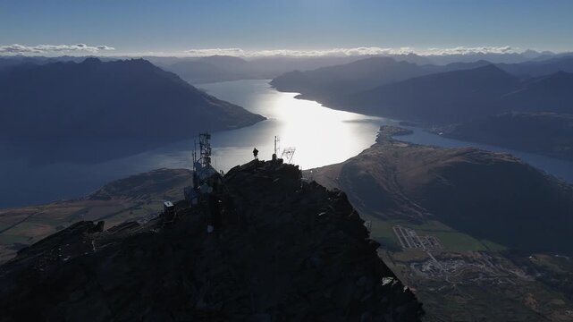 Aerial silhouette of man at comm tower on mtn peak above silvery lake