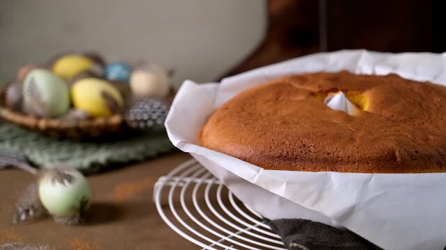 Freshly baked Portuguese sponge cake rests on a wire rack in its paper lining. Soft natural light highlights the airy texture and rustic holiday setting with colorful eggs in the background.