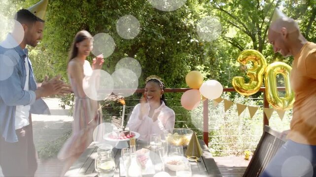 Seated honoree reacting guest bringing lit cake event overlays floating gold30 balloon around her