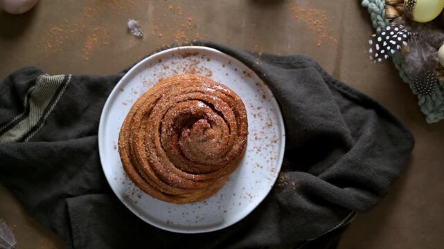 Top view of a glazed cinnamon bun dusted with spice on a ceramic plate. Soft, moody lighting highlights the flaky pastry texture against dark linens and seasonal Easter egg accents.