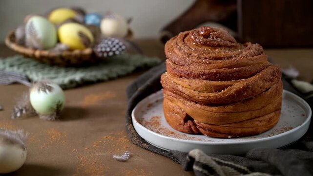 Large spiral cinnamon bun dusted with sugar on a white plate. The background features colorful Easter eggs in a basket and soft feathers, creating a warm festive atmosphere with shallow depth of field