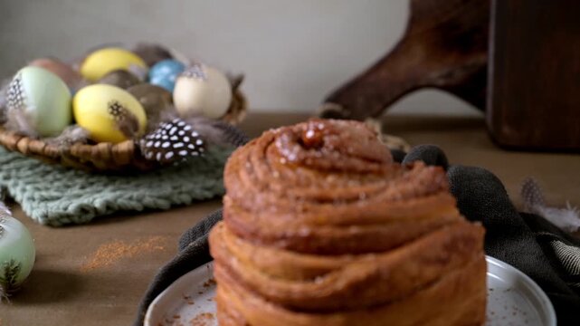 A golden-brown cruffin dusted with sugar and cinnamon sits on a ceramic plate. Soft natural lighting highlights the flaky pastry layers against a rustic background with colorful Easter eggs.
