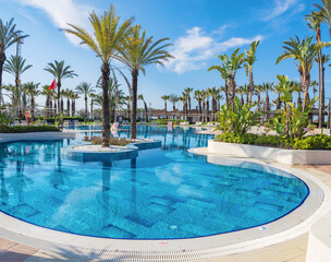 Fototapeta premium Wide angle view of a stunning leisure area in a tropical hotel resort with elegant pool design, surrounded by sun loungers and palm trees under a bright summer sun, idyllic holiday in Turkey.