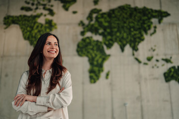 Confident global business woman standing with arms crossed next to a green plant world map for sustainability and growth © Zamrznuti tonovi