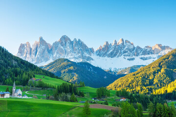 Fototapeta premium Santa Magdalena church in Val di Funes valley with alpine village, green hills and Odle snow peaks in Dolomites mountains at sunrise, South Tyrol, Italy. Rural landscape in Italian Dolomiti
