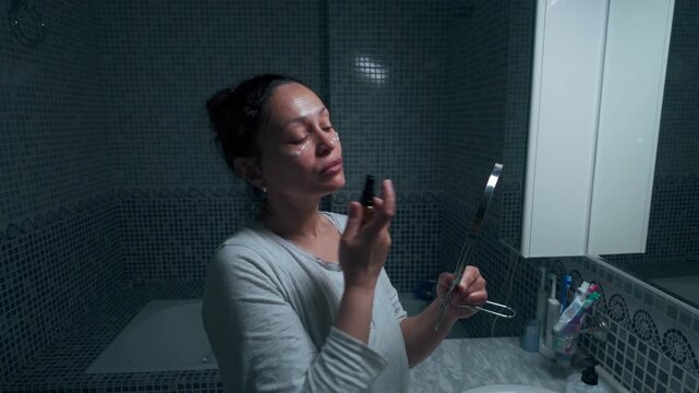 Daily routine of a woman applying eye cream in a bathroom mirror, holding a small mirror and makeup applicator during a quiet skincare moment.