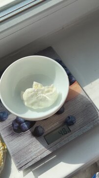 tablespoon of yogurt or sour cream is weighed to 50 grams on a white plate, showing precise portioning step-by-step for healthy dessert, breakfast, or culinary preparation.