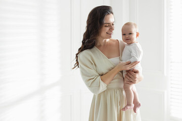 Smiling mother in dress holding her cute baby near white wall at home. Space for text © New Africa