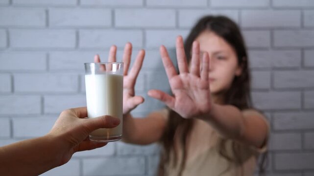 Girl refusing to drink a glass of milk. Unhappy young girl gesturing with her hands to refuse a glass of milk offered by an adult, expressing a clear sign of rejection due to lactose intolerance