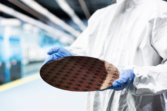 Holding processed silicon wafer, cleanroom and technician in automated fabrication plant. Engineer inspecting semiconductor substrate, defects and engineer doing quality control on microelectronics
