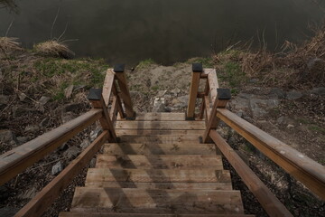 Naklejka premium Top-down perspective of a wooden staircase leading to a riverbank. High-angle view showing timber steps, railings with black caps, and rocky ground near dark water.