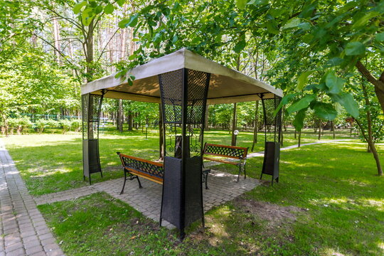 People enjoy sitting on benches under a gazebo in a park. The area is surrounded by green trees. It is sunny and they relax in the afternoon shade