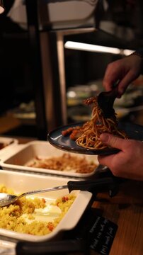 Vertical screen close up person serving noodles at buffet table soft dim lighting warm atmosphere food service concept self service dining and hospitality scene