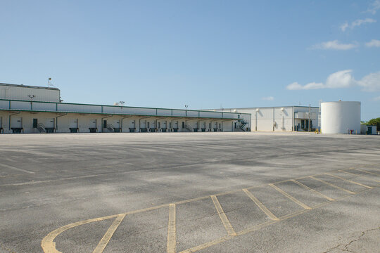 Large industrial warehouse with multiple loading docks, empty asphalt yard and stair access under a clear blue sky, conveying logistics, transport infrastructure, storage and quiet commercial space.
