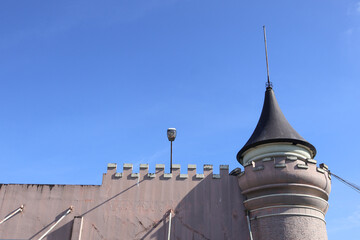 ​An old, weathered castle wall with battlements and a round turret topped by a conical black spire stands against a clear, deep blue sky during daytime, showing architectural detail. © Jumain