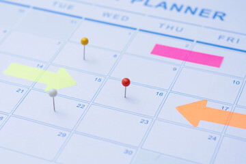 Close up of a monthly wall calendar featuring push pins and bright arrow post-it notes to mark...
