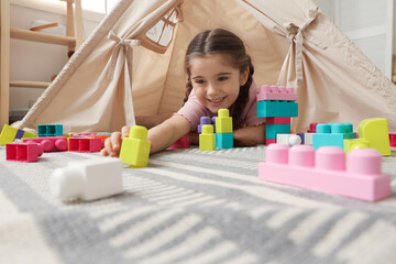 Little girl playing with building blocks near toy wigwam in playroom, low angle view © New Africa