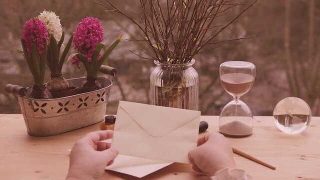 Female Hands holding blank beige envelope on wooden table, pink hyacinth and willow branches in glass jar, ink bottles, Easter spring atmosphere, writing letter, vintage stationery, copy space