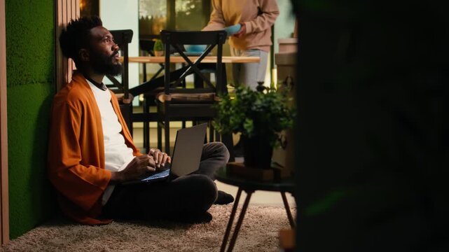 Cozy lifestyle scene of African American man relaxing indoors with his laptop, browsing websites online and doomscrolling. Person surrounded by warm light and comfortable home ambiance.