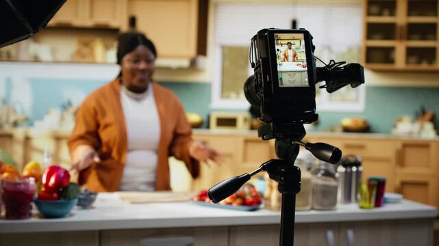 Behind the scenes of black woman recording meal prep vlog with tripod in the kitchen, presenting cooking process for online social media audience. Female influencer and content creator.