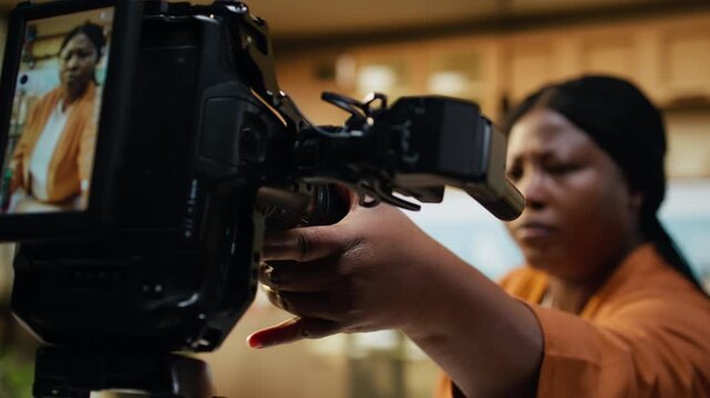 Behind the scenes of African American vlogger adjusting camera on tripod in home kitchen studio. Content creator checks lighting settings and steady framing during preparations, ready for filming.