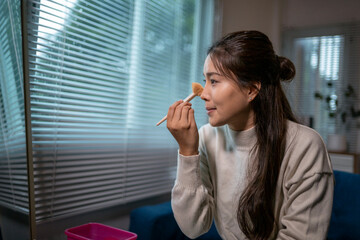 Fototapeta premium Young asian woman applying makeup with brush near window blinds