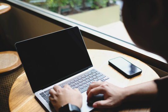 An over the shoulder view of a person typing on a laptop with a blank black screen placed on a wooden table in a cafe.