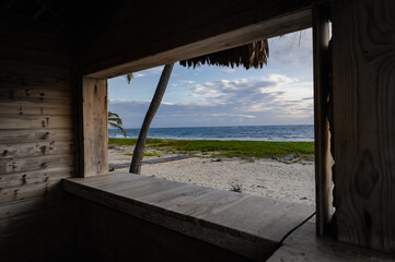 Obraz premium A stunning seaside view framed by a rustic wooden window, showcasing serene waves, lush green grass, and swaying palm trees under a beautiful sky at twilight.