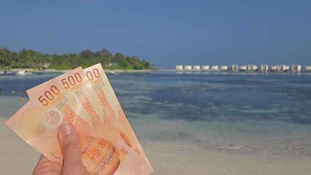Stack of 500 maldivian ruffiyaa Banknotes in hand with maldivian island and ocean in background. Expencive travel concept