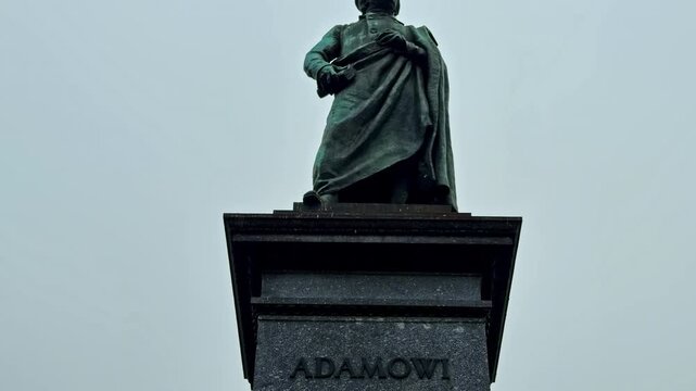 Pan down on the Adam Mickiewicz Monument in Krakow, Poland, from the bronze figure to the engraved pedestal and allegorical sculptures under an overcast sky.