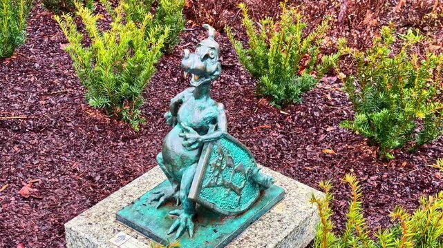 Small bronze dragon statue on a stone base in a garden bed near Corpus Christi Basilica in Krakow, Poland, surrounded by shrubs and wet mulch.