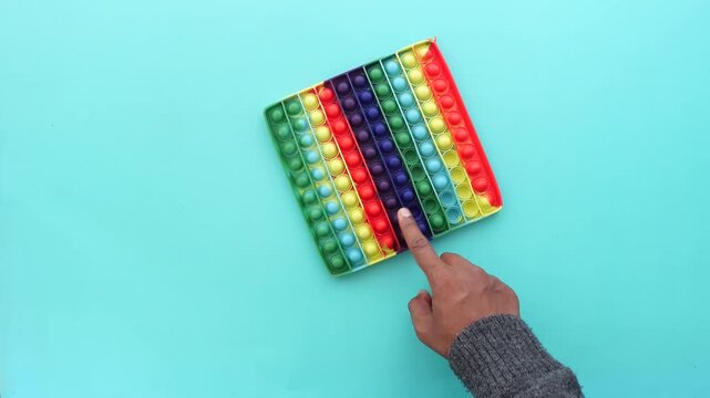 Child's hand pushing colorful pop it fidget toy on blue background top view, motion clip.