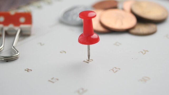 Person pinning red pushpin on sixteenth day of calendar month over coins and binder clip.
