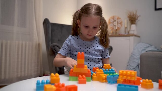 Cute Caucasian five year old girl playing with plastic blocks at home on white glass table, assembling cubes 