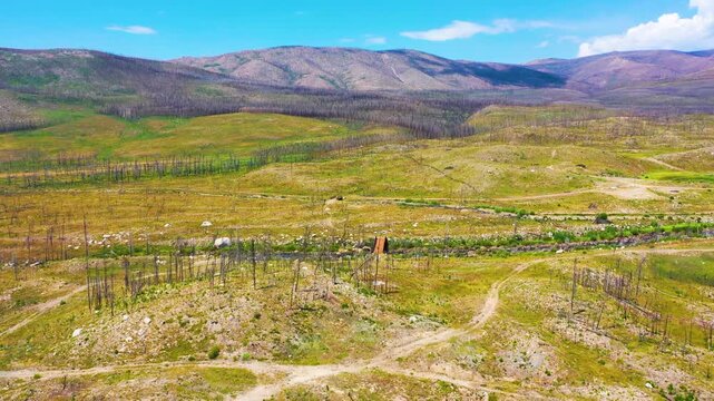 Aerial panorama of a burned mountain landscape near Grand Lake Colorado, with winding dirt paths, exposed soil, recovering grassland, and broad ridgelines under blue sky.