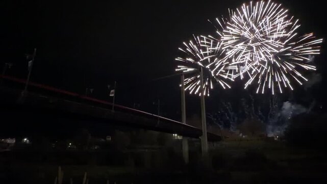 Fireworks illuminating the night sky over a bridge and river, creating colorful reflections during the traditional Fallas de Valencia festival