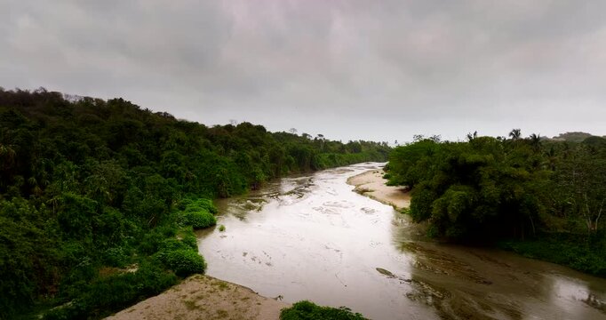 Dolly forward aerial shot above Rio Piedras in Los Naranjos, Colombia, following a muddy tropical river between dense rainforest banks under a grey overcast sky