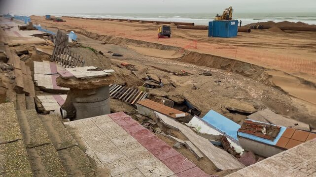 Coastal promenade showing severe erosion and structural damage after a powerful storm, with ongoing beach reconstruction efforts