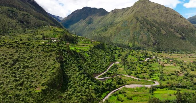 Rural road winding through the green terraced fields of a valley in the Andes near Ollantaytambo, Peru. Aerial drone panoramic view