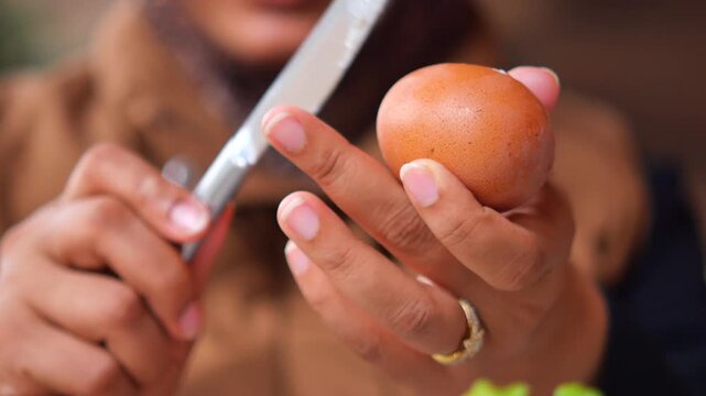 Woman hands peeling boiled chicken egg shell with butter knife in outdoor kitchen setting.