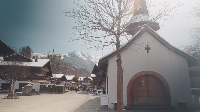 Saint Nicholas chapel with main street, Gstaad Palace and snowy mountains in Canton of Bern, Switzerland, wide shot, static