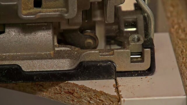Slow motion handheld close up of a power jigsaw cutting a laminated chipboard panel, saw blade slicing a wood board with dust on workshop table during DIY carpentry and renovation