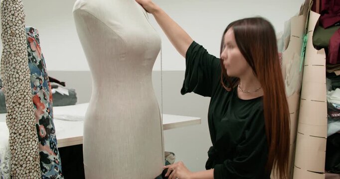 Designer workshop. Taking measurements. Female seamstress inspecting size of tailor dummy mannequin with tape in room with fabric roll.